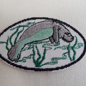 May include: Embroidered patch of a gray and green manatee swimming in a bed of green seaweed. The patch is oval shaped and has a black outline.