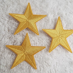 Puede incluir: Tres parches dorados en forma de estrella sobre una superficie blanca y peluda. Las estrellas están bordadas con un diseño detallado.