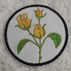 May include: A round embroidered patch with a black border featuring three yellow roses with green leaves and stems on a white background.