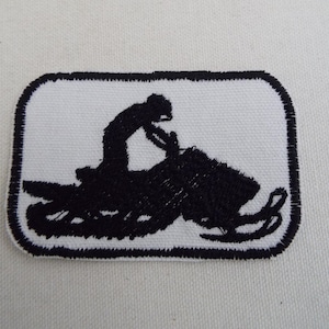 May include: Black and white embroidered patch of a person riding a snowmobile. The patch is rectangular and has a white background with a black outline.