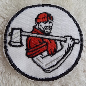 Op de afbeelding: Een witte geborduurde patch met een zwarte rand, met een cartoon-houtvester met een rode muts en shirt, die een bijl vasthoudt.