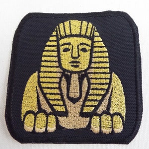 May include: A black fabric patch with an embroidered image of a golden pharaoh wearing a headdress. The pharaoh is sitting with its arms crossed and has a serious expression.