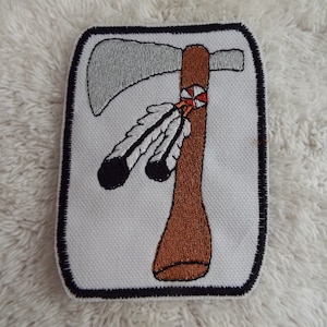 Puede incluir: Un parche bordado blanco con un contorno negro que representa un hacha marrón con una hoja plateada y dos plumas negras y blancas.
