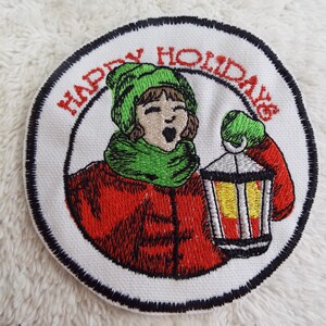 Op de afbeelding: Een witte geborduurde patch met een zwarte rand. De patch toont een persoon die een rode jas, een groene hoed en een sjaal draagt en een lantaarn vasthoudt. De tekst "Happy Holidays" is rond de persoon geborduurd.