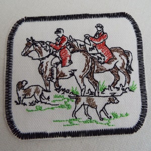 May include: Embroidered patch featuring two people riding horses, with a brown and white dog in the foreground. The patch is surrounded by a black border.