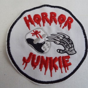 May include: A white embroidered patch with a black outline. The patch features a red dripping text that says "HORROR JUNKIE". A gray hand with claws is reaching for a white skull with red blood dripping from it.