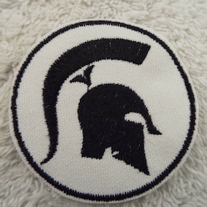 Könnte beinhalten: Ein weißer runder Patch mit einem schwarzen gestickten Spartanerhelm.