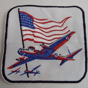 May include: Embroidered patch featuring a blue and red airplane flying over a waving American flag. Three smaller airplanes are flying below the larger plane.