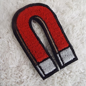 May include: Embroidered patch in the shape of a horseshoe magnet. The magnet is red with a black border and silver tips. The patch is on a white, textured surface.