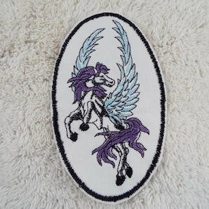 Puede incluir: Un parche bordado con un caballo blanco alado con crin y cola morados, sobre un fondo ovalado blanco con contorno negro.