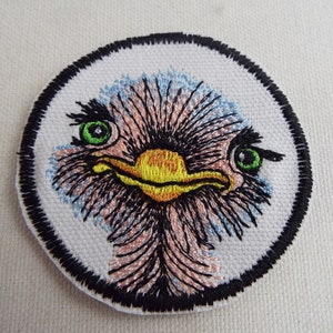 May include: Embroidered patch of an ostrich's face with a black outline, green eyes, and a yellow beak. The ostrich's feathers are pink and blue.
