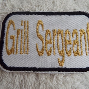 Puede incluir: Parche blanco bordado con borde negro, con el texto "Gill Sergeant" en hilo dorado.