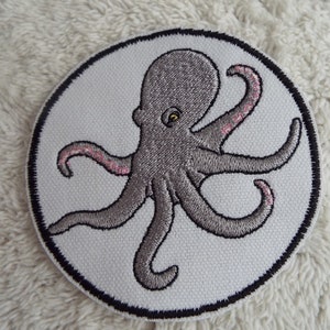 Puede incluir: Un pulpo bordado gris con tentáculos rosados ​​en un parche circular blanco con un borde negro.
