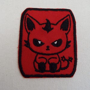 Puede incluir: Parche rojo bordado para planchar con un gato de dibujos animados con un cuerno de diablo, ojos negros y un contorno negro. El gato está sentado con los brazos cruzados.