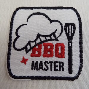 Może przedstawiać: Biała naszywka haftowana z czarnym obramowaniem. Naszywka przedstawia czapkę szefa kuchni, haftowany czerwony tekst "BBQ Master" i czarny szpatułkę.