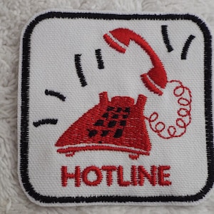 May include: Red embroidered patch with a white background and black border. The patch features a red rotary phone with a black dial and a red handset. The phone has a red curly cord. The text "HOTLINE" is embroidered in red below the phone.