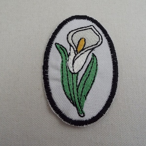 Könnte beinhalten: Ovaler, gestickter Aufnäher mit einer Calla-Lilie. Die Lilie hat eine weiße Blüte mit gelbem Blütenkolben, grüne Blätter und ist vor einem weißen Hintergrund platziert. Der Aufnäher ist mit schwarzem Garn umrandet.