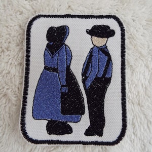 May include: Embroidered patch featuring a couple in traditional Amish clothing. The woman is wearing a blue dress and black bonnet, while the man is wearing a blue shirt, black pants, and a black hat.