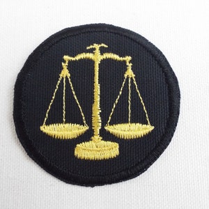 Könnte beinhalten: Ein schwarzer runder Patch mit einem goldenen gestickten Justitia-Symbol.