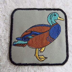 May include: A brown, blue, and green embroidered patch of a duck with a black border.
