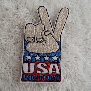 Puede incluir: Parche bordado con un gesto de mano en señal de paz en beige, azul, rojo y blanco. La mano lleva una manga a rayas azul y blanca con estrellas blancas. El texto "USA VICTORY" está escrito en rojo y blanco.