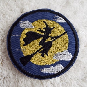 May include: Embroidered patch featuring a black silhouette of a witch flying on a broomstick in front of a yellow moon with white clouds on a blue background.