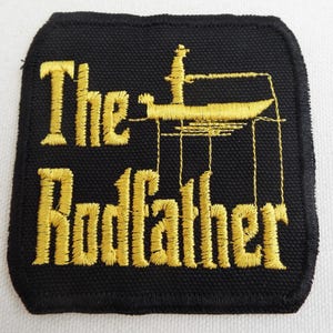 Może przedstawiać: Czarna kwadratowa naszywka ze złotym haftowanym tekstem i wzorem. Tekst brzmi "The Rodfather" z grafiką wędki i kołowrotka. Naszywka ma czarną obwódkę i prawdopodobnie jest naszywką do naprasowywania lub przyszywania.