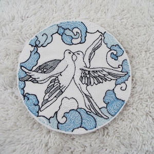 Puede incluir: Dos palomas blancas bordadas sobre un fondo blanco con nubes azules. Las palomas se miran y parecen besarse. Las nubes son estilizadas y tienen un diseño en espiral.