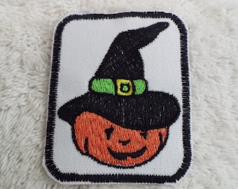 May include: A white fabric patch with a black outline, featuring an orange pumpkin with a black witch's hat. The hat has a green band with a yellow buckle.