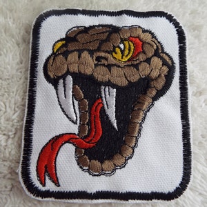 May include: Embroidered patch featuring a detailed snake head with an open mouth, fangs, and a red tongue. The snake has brown and black scales, yellow eyes, and is set against a white background with a black border. Ideal for jackets or bags.