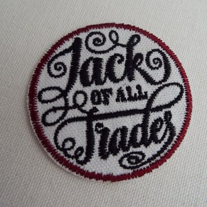 Op de afbeelding: Een witte geborduurde patch met een rode rand. De patch heeft de tekst "Jack of all Trades" in een gestileerd zwart lettertype.