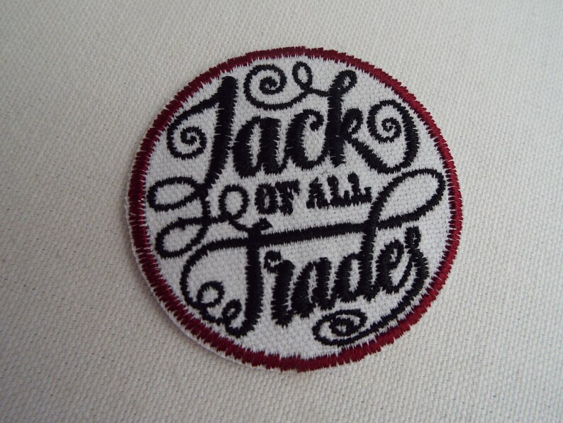 Op de afbeelding: Een witte geborduurde patch met een rode rand. De patch heeft de tekst "Jack of all Trades" in een gestileerd zwart lettertype.