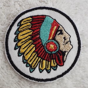May include: Embroidered patch featuring a Native American man in profile wearing a colorful headdress with red, yellow, and turquoise feathers. The patch is circular with a black border and white background.