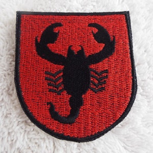 May include: A red embroidered patch with a black scorpion design. The scorpion is facing forward with its claws raised. The patch is shaped like a shield.