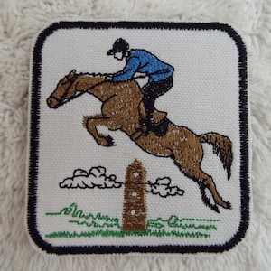 May include: Embroidered patch of a brown horse jumping over a fence with a rider in a blue jacket and white helmet.
