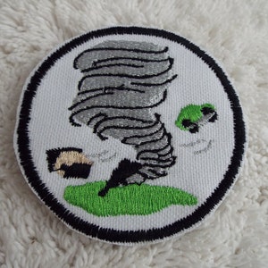 May include: A white embroidered patch with a black border depicting a gray tornado with a green car being lifted into the air.