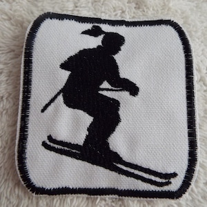 May include: Black and white embroidered patch of a skier in a square shape with a black border.