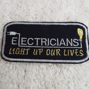 May include: Black embroidered patch with white stitching. The patch says "Electricians Light Up Our Lives" with a lightbulb and electrical cord graphic.