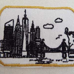 Puede incluir: Parche bordado con un horizonte de ciudad en blanco y negro, que incluye edificios altos y un puente. El parche tiene un fondo blanco con un borde dorado. Dos nubes y un pájaro también están incluidos en el diseño.