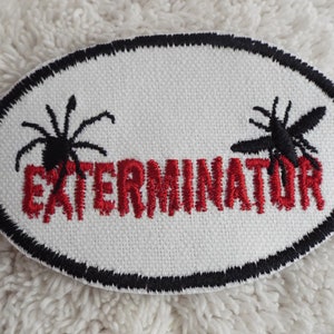 May include: White oval patch with black embroidered border. The patch features a red embroidered word "Exterminator" with a black embroidered spider and fly on either side.