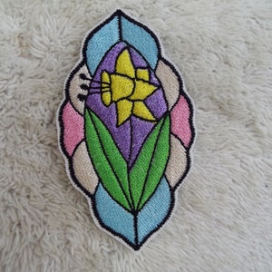 May include: A colorful embroidered patch featuring a yellow daffodil with green leaves, surrounded by a stained glass-like design in shades of pink, blue, and purple.
