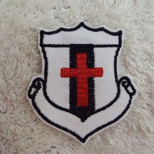 Puede incluir: Parche blanco bordado con un contorno negro en forma de escudo con una cruz roja en el centro.