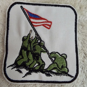 May include: An embroidered patch depicting the iconic image of the raising of the American flag on Iwo Jima. Five soldiers in green uniforms are raising the flag on a hilltop. The flag is red, white, and blue.