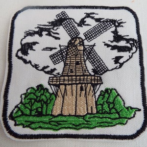May include: Embroidered patch featuring a brown windmill with black blades against a white background. The windmill is surrounded by green trees and a white cloud.