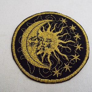 Puede incluir: Un parche redondo bordado de color negro con un diseño de sol y luna dorado. El sol tiene una cara y rayos, y la luna es una media luna. Hay pequeñas estrellas alrededor del sol y la luna.