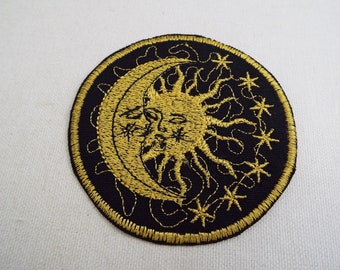 May include: A round, black embroidered patch featuring a gold sun and moon design. The sun has a face and rays, and the moon is a crescent shape. There are small stars surrounding the sun and moon.