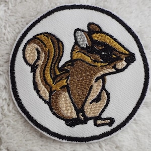 May include: A white embroidered patch with a black outline, featuring a brown and tan cartoon squirrel with a bushy tail.