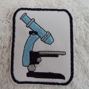 Puede incluir: Parche bordado con un diseño de microscopio azul claro y negro sobre fondo blanco. El parche tiene un borde negro y es rectangular con esquinas redondeadas. Ideal para entusiastas de la ciencia.