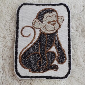 Könnte beinhalten: Ein gestickter Aufnäher mit einem braunen und schwarzen Affen auf weißem Hintergrund. Der Affe sitzt mit seinem Schwanz um seinen Körper gewickelt.