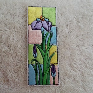 May include: An embroidered patch featuring a purple iris flower with green leaves and buds, set against a colorful stained glass background.